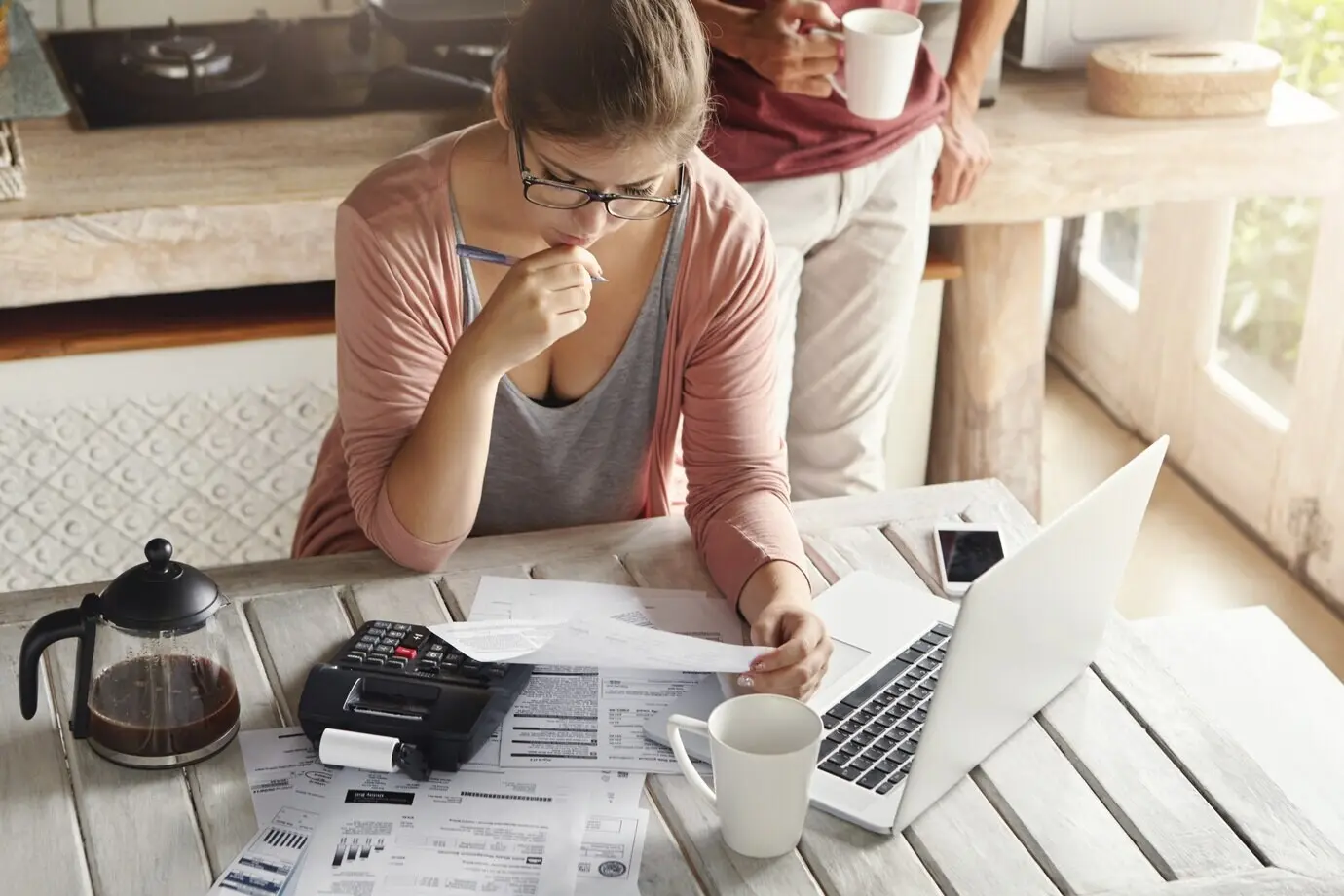 A couple struggling with debt and unable to pay off their mortgage. A thoughtful, frustrated-looking woman holds a pen while managing the family budget, calculating figures with a calculator and a notebook PC.
