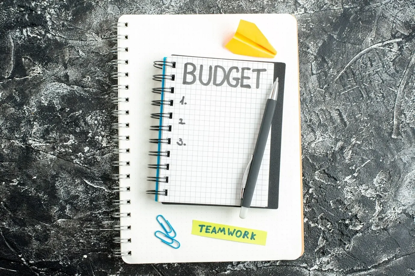 Top view of a notepad with a note reading “budget” and a pen on a gray background, with a colored student copybook—school, college, dark, business, money, lessons.